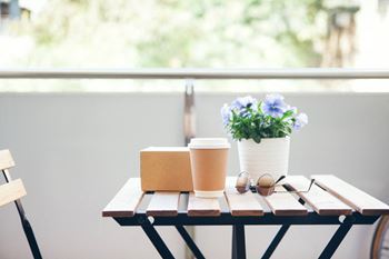 Outdoor patio area with table and chairs.  Table has a take out container, pair of sunglasses, potted plant and Paper Coffee Cup.at Fountains of Fair Oaks, California, 95628